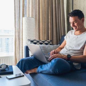 young attractive man sitting on sofa at home working on laptop online, using internet, smiling, happy mood, freelancer, free leisure time, relaxed, modern job lifestyle