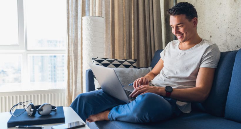 young attractive man sitting on sofa at home working on laptop online, using internet, smiling, happy mood, freelancer, free leisure time, relaxed, modern job lifestyle