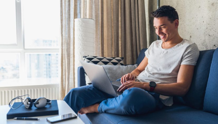 young attractive man sitting on sofa at home working on laptop online, using internet, smiling, happy mood, freelancer, free leisure time, relaxed, modern job lifestyle